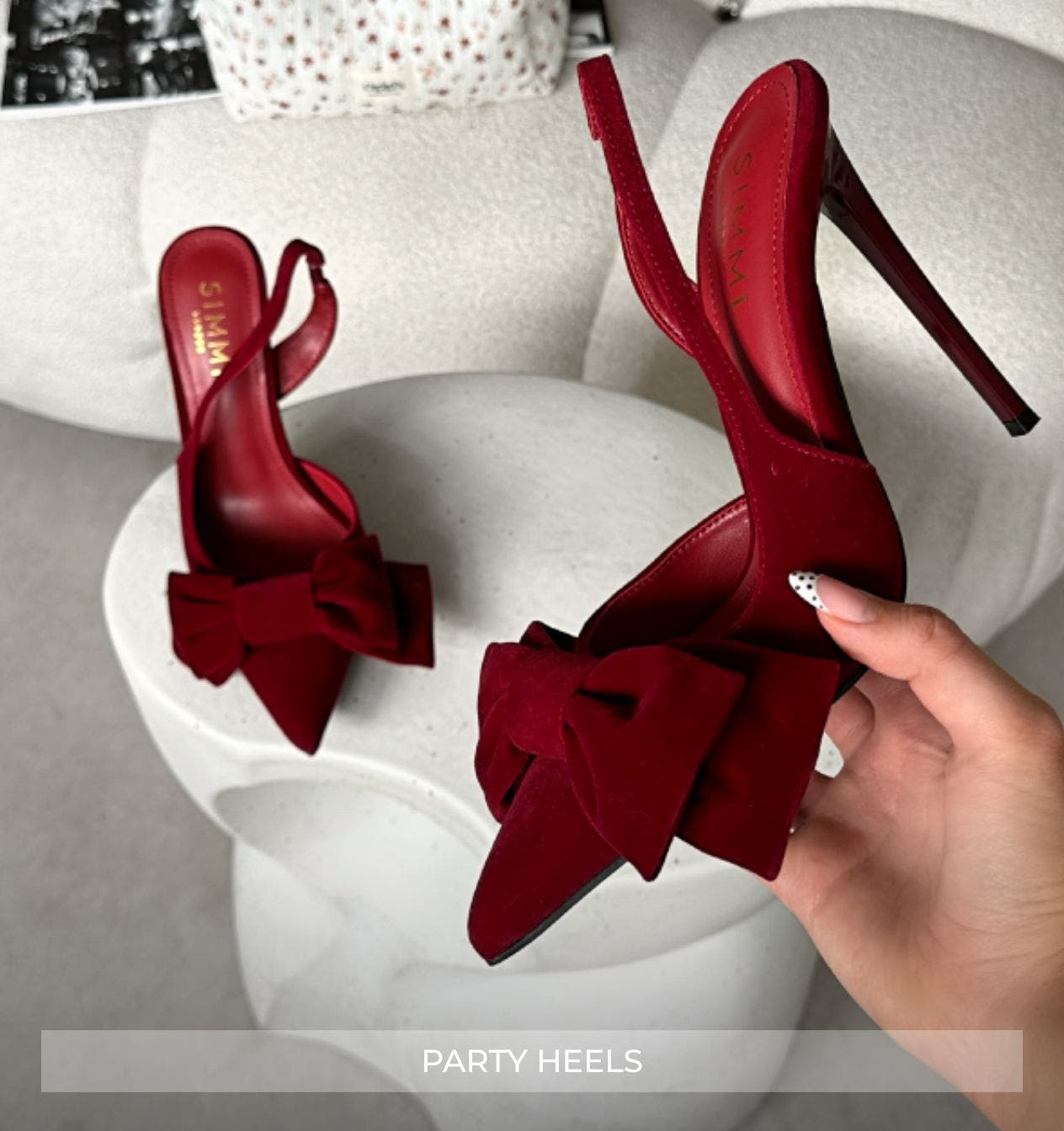 Red high-heeled shoes with bows held by a hand on a white surface.