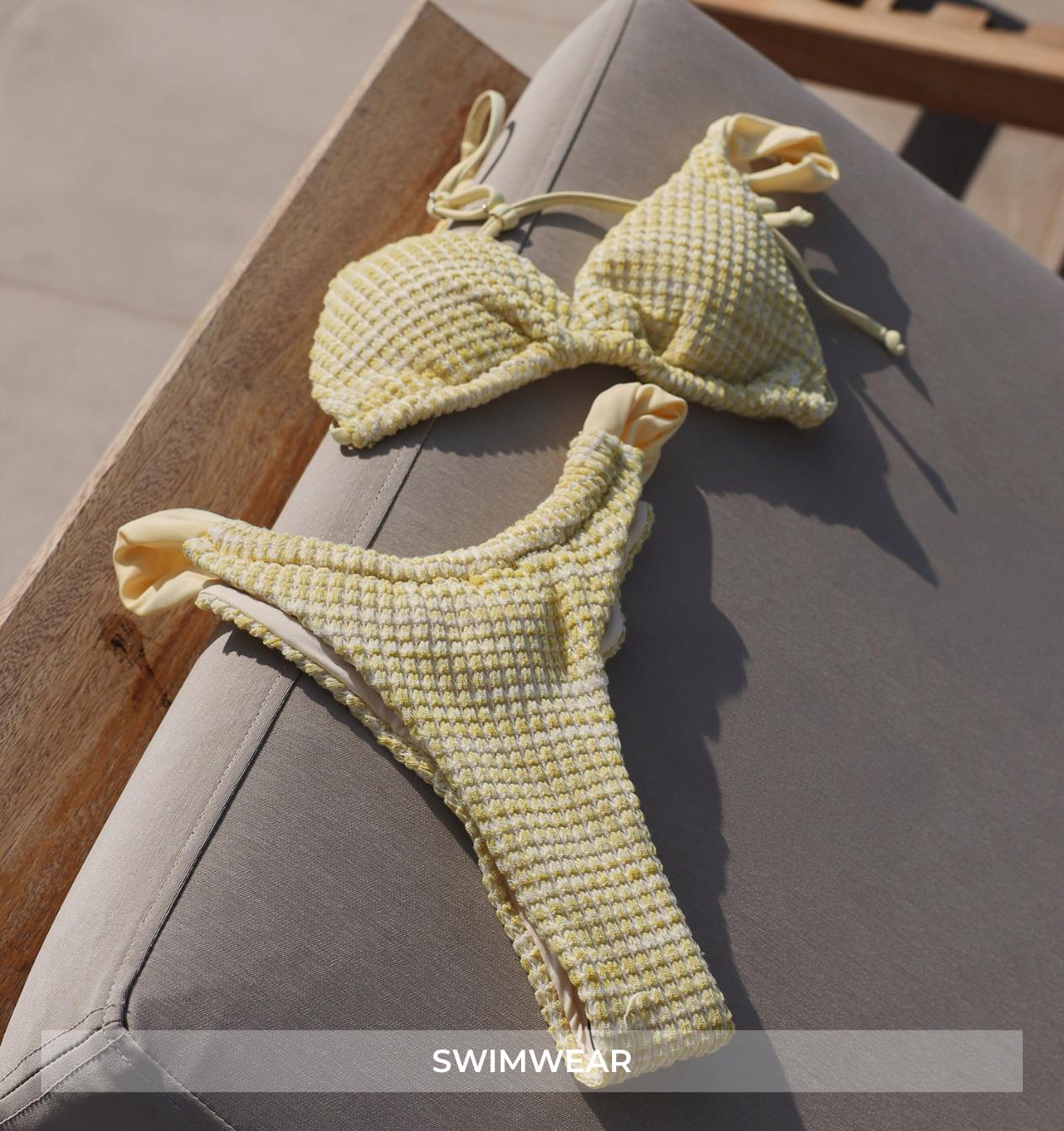 A pale yellow textured bikini set laid on a gray cushioned lounge chair outdoors. The top has braided cups with tie details, and the bottoms are thong-style with a textured woven pattern.