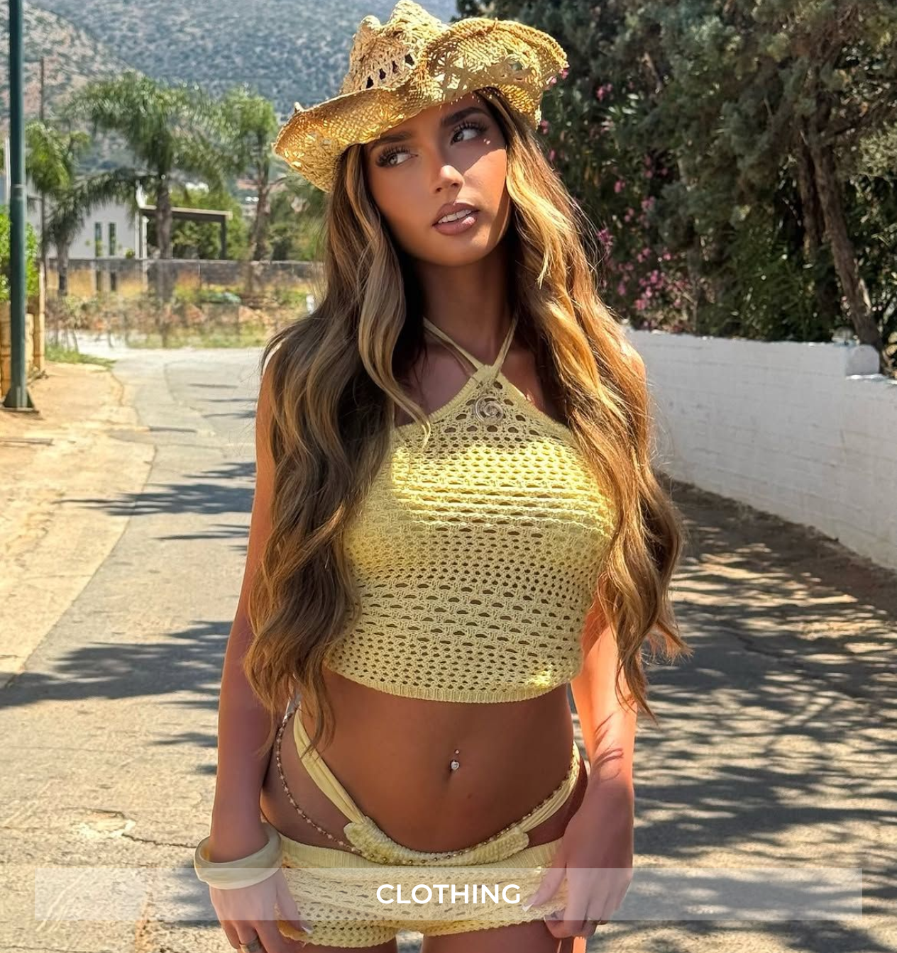 A woman wearing a light yellow crochet knit two-piece set consisting of a halter crop top and matching strappy bikini-style bottoms, along with a straw cowboy hat, standing outdoors on a sunlit road.