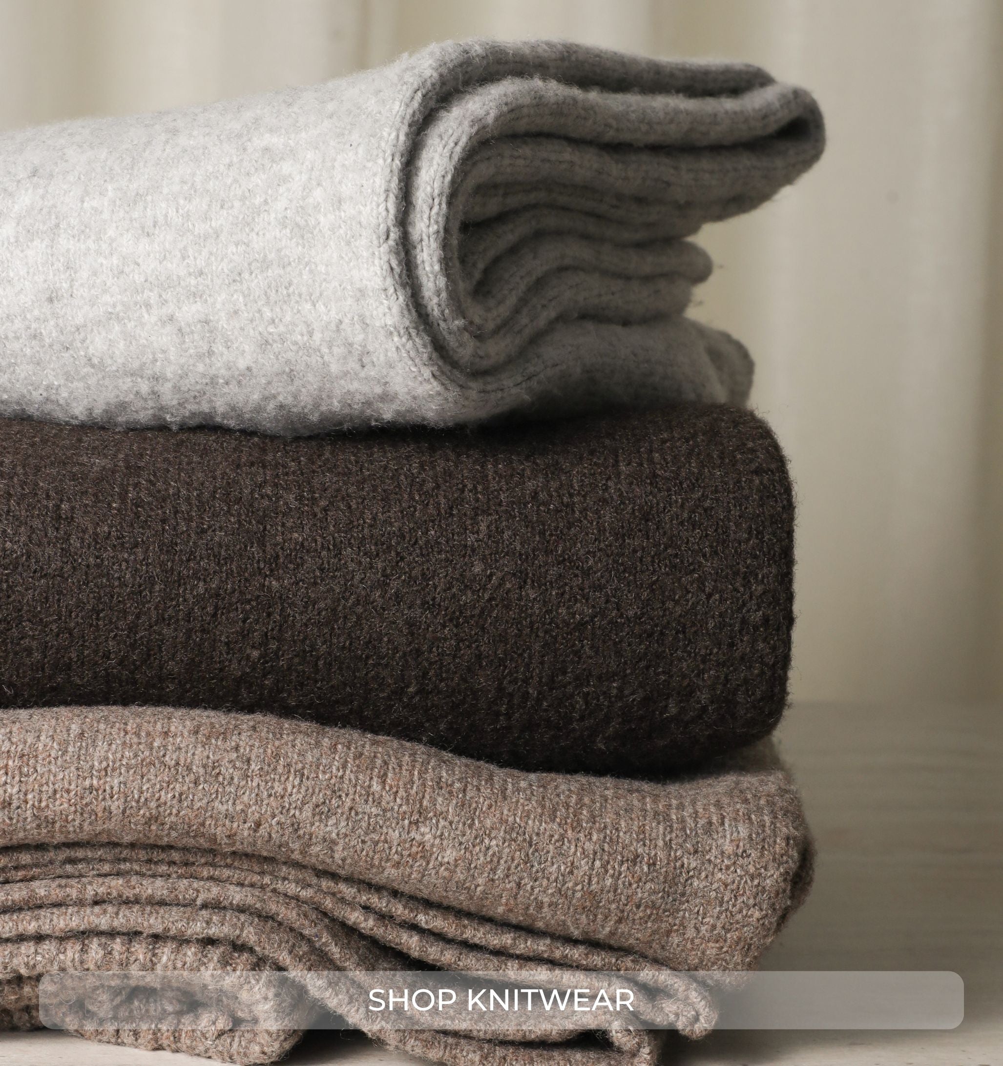 Stack of folded knitwear in gray, brown, and beige on a neutral background with 'Shop Knitwear' text.