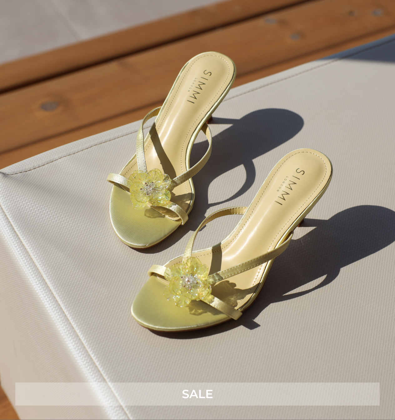 A pair of pale yellow heeled thong sandals with floral embellishments and rhinestone centers, displayed on a light-colored textured surface with a wooden bench in the background.