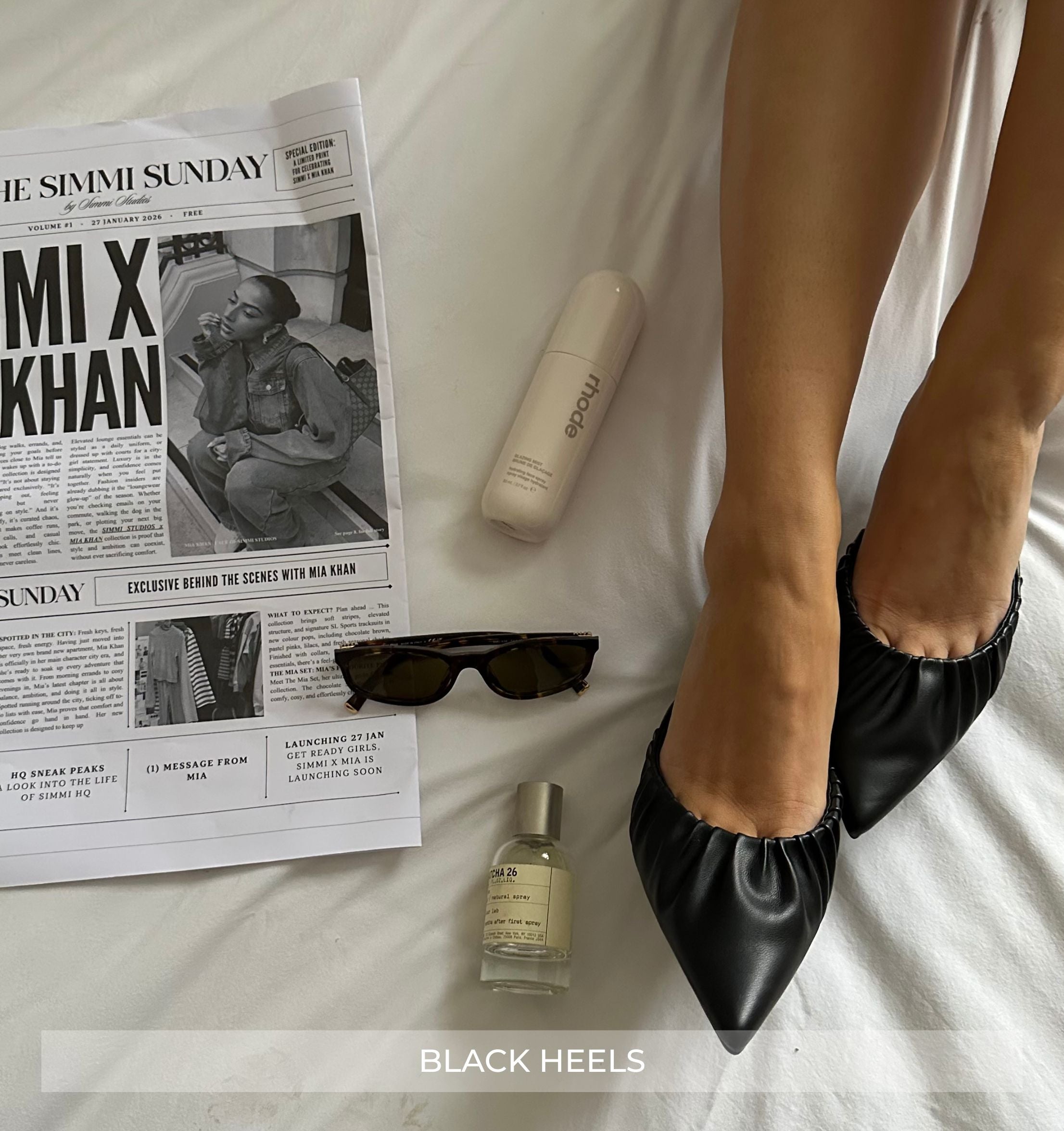 Black heels on a bed with a newspaper, bottle, and sunglasses.