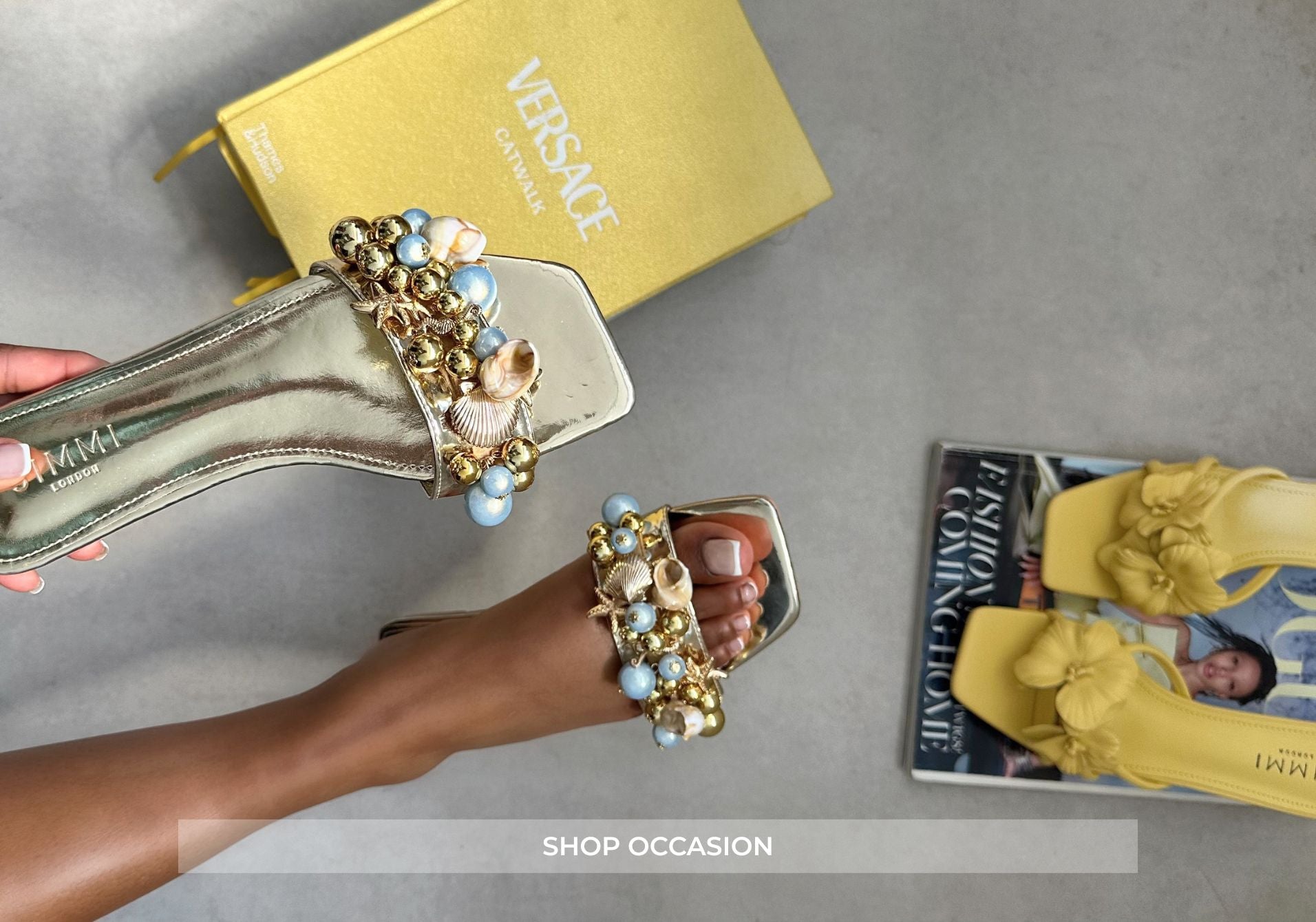 Decorative high-heeled shoes with embellishments held by a hand, Versace box in the background.