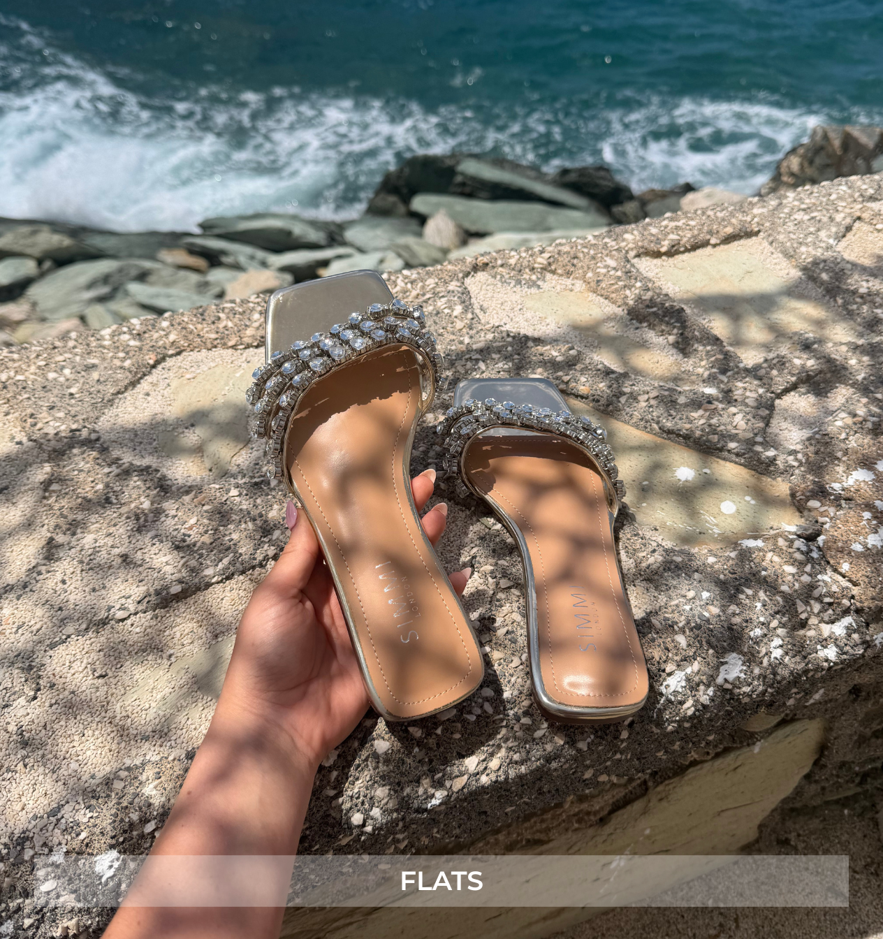 Pair of embellished sandals held over a rocky surface with ocean view