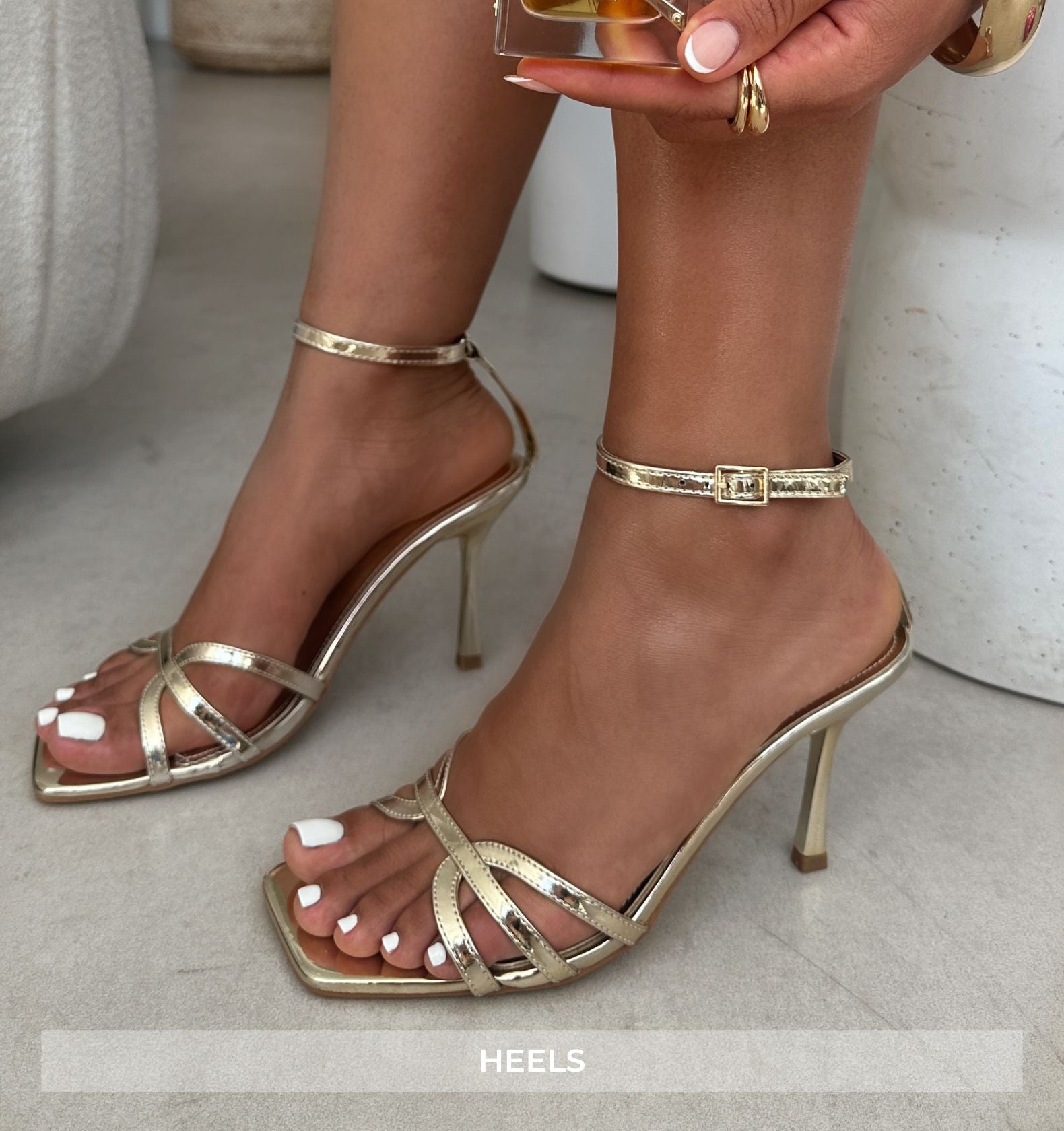 Gold high-heeled sandals worn by a person on a light gray floor.