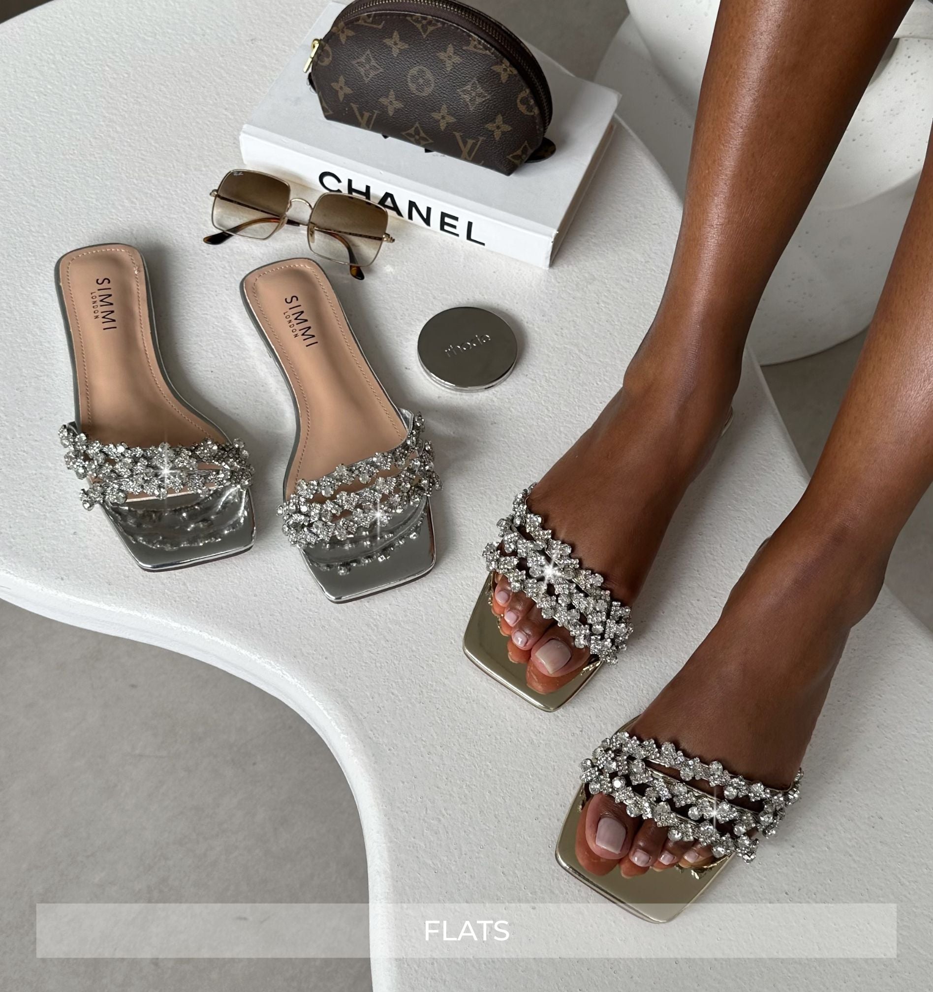 Elevated view of embellished sandals with a Chanel box and sunglasses on a white surface.