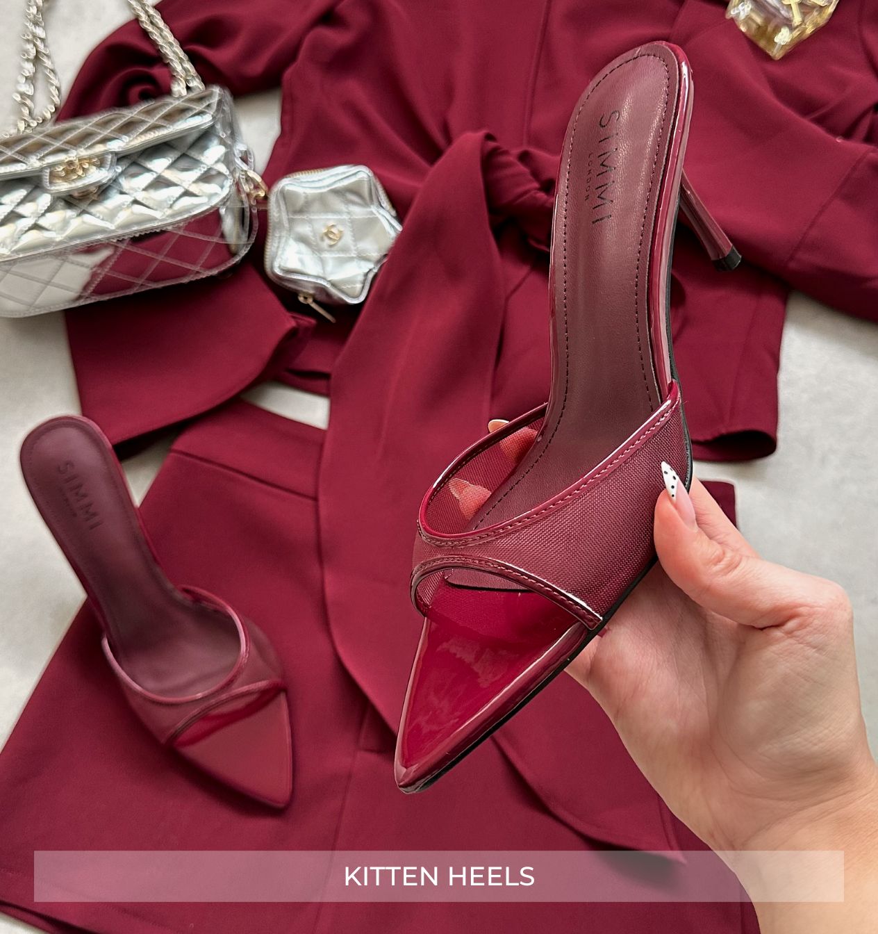 Red high-heeled shoe held by a hand with a red fabric and silver purse in the background.