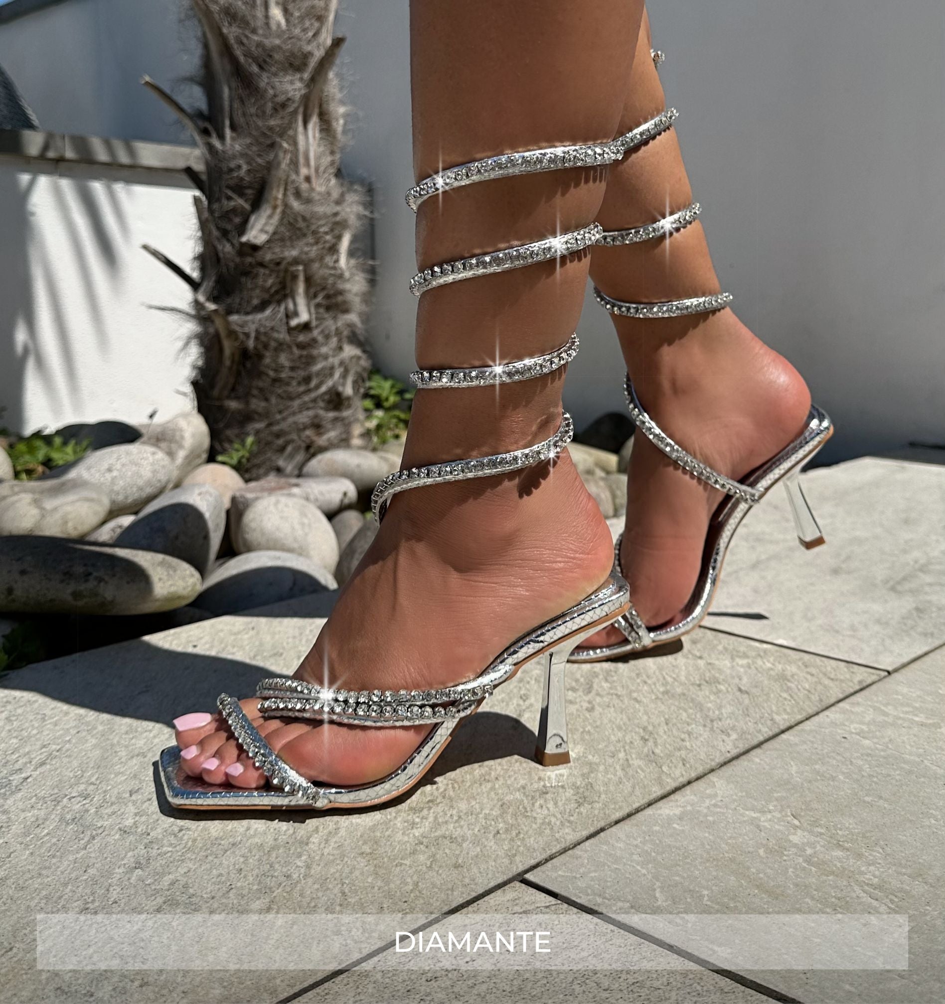 Silver strappy high-heeled sandals on a stone surface with 'DIAMANTE' branding.