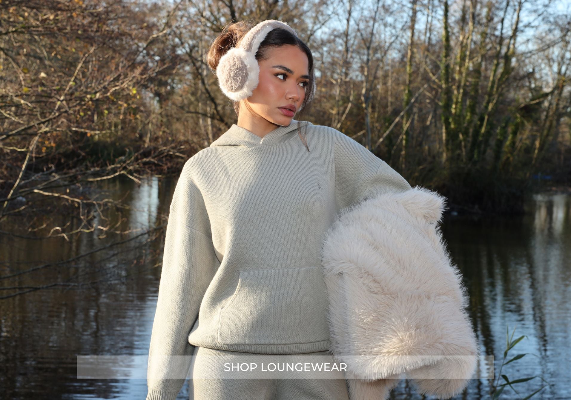 Woman wearing a light gray hoodie and white ear muffs by a body of water.