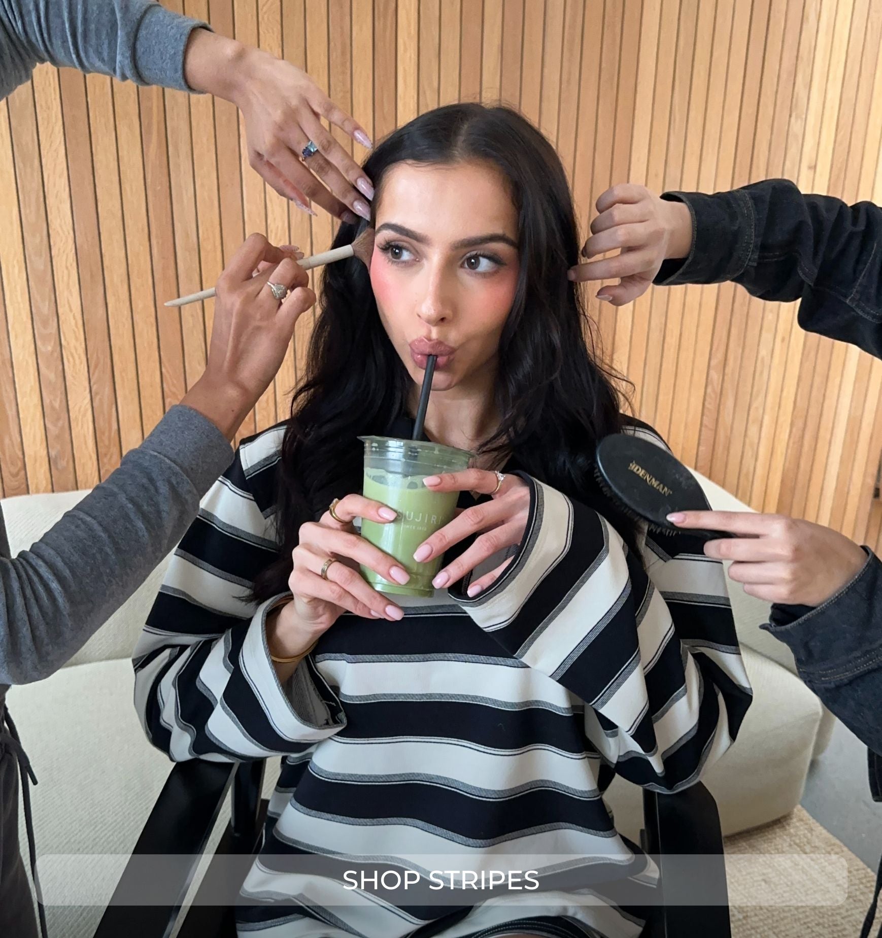 Woman in a striped sweater holding a drink, surrounded by people applying makeup and hair products.