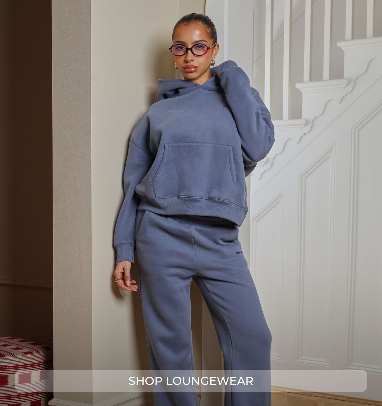 Woman wearing a blue tracksuit standing in a home setting with text 'Shop Loungewear'.