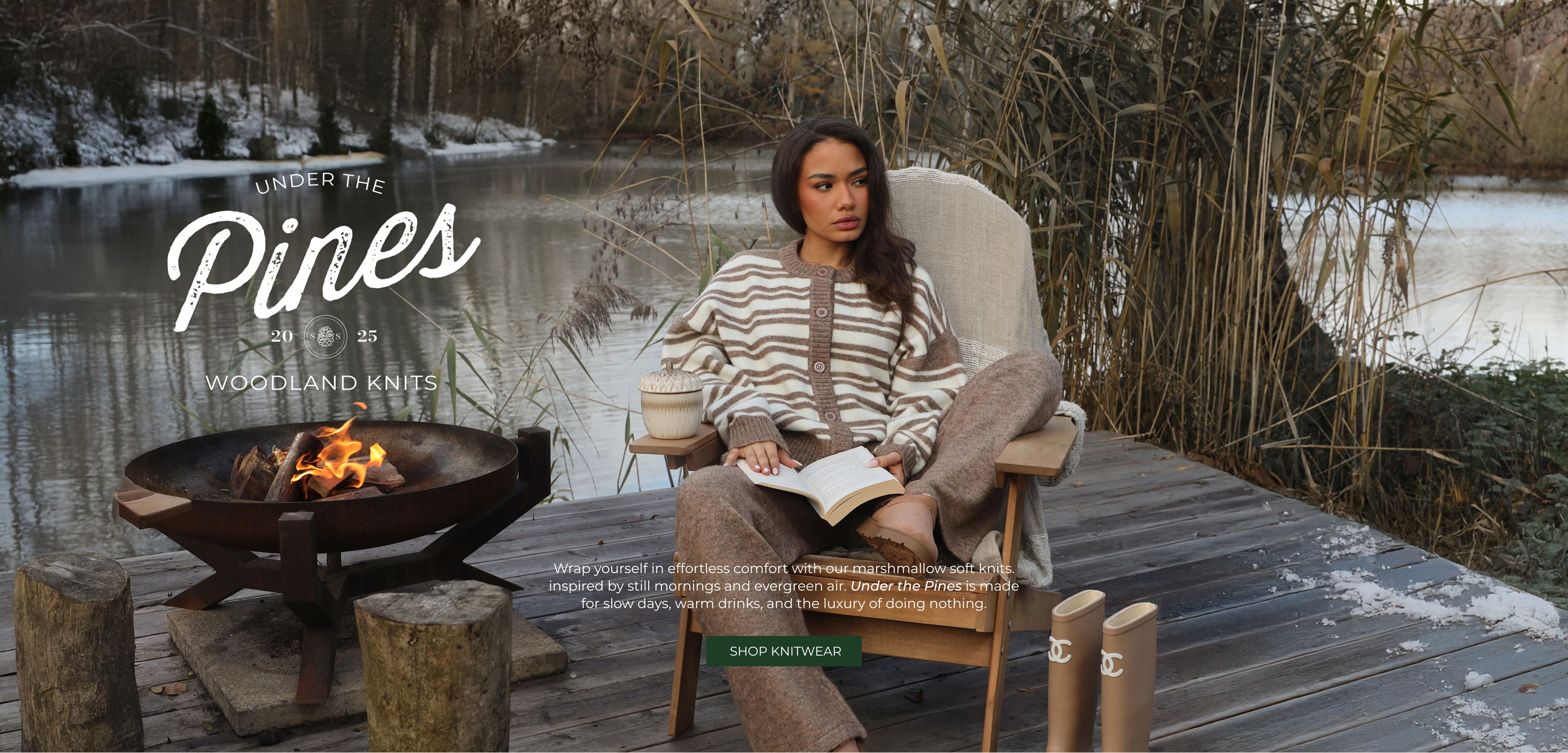 Woman sitting on a wooden deck by a lake with a fire pit, surrounded by nature.